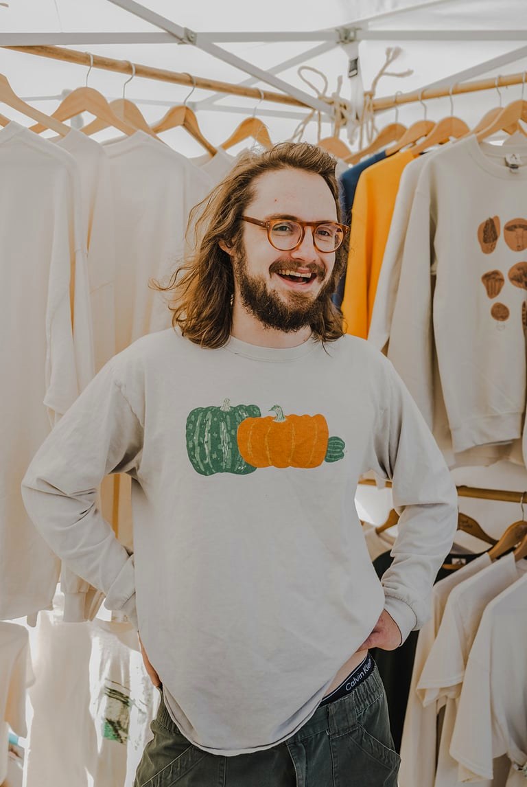 A smiling vendor in a pumpkin sweatshirt in front of his rack of printed shirts